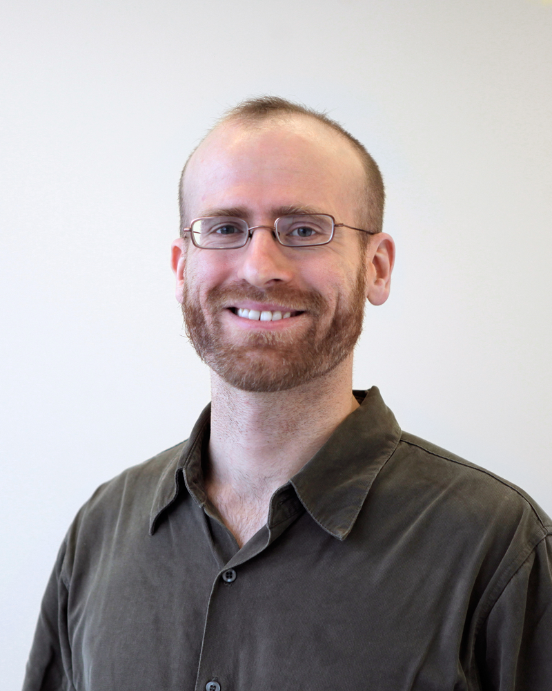 Portrait headshot of Anselm Bradford, who is wearing wire glasses and a dark gray collared shirt.