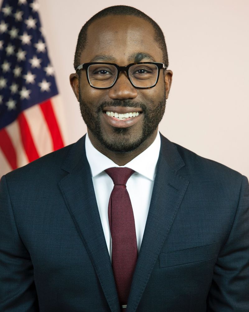 In the foreground, Brian Whittaker smiles, wearing dark glasses, a dark gray suit, white shirt, and maroon tie. He has a beard and moustache. In the background is a light colored wall and the American flag stands behind him on his right.