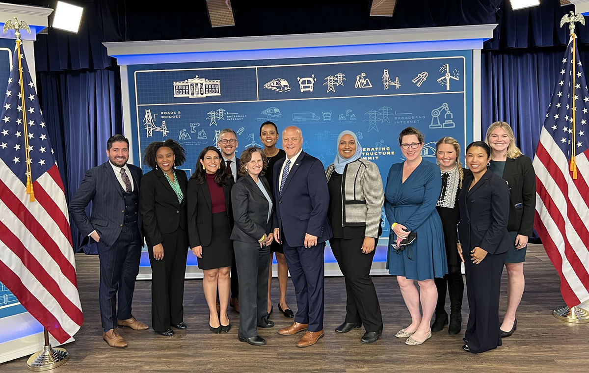 U.S. Digital Corps Fellow Meredith Brown, right, poses on stage with 11 members of the White House Infrastructure Implementation team. Attendees are smiling in business clothing and are standing between two American flags.