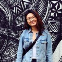 
Jenny Wang stands in front of a large black and white piece of art. She is smiling, and is wearing glasses, a white shirt and light blue denim jacket.