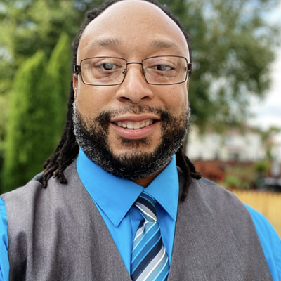 
Joe Lindsey stands outside near trees. He is wearing a gray vest, light blue shirt, and striped tie.